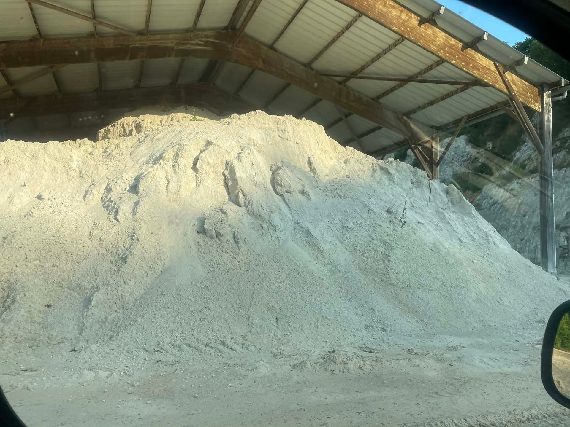 Un grand tas conique de sable ou de gravier de couleur claire, stocké sous un abri à toit métallique.