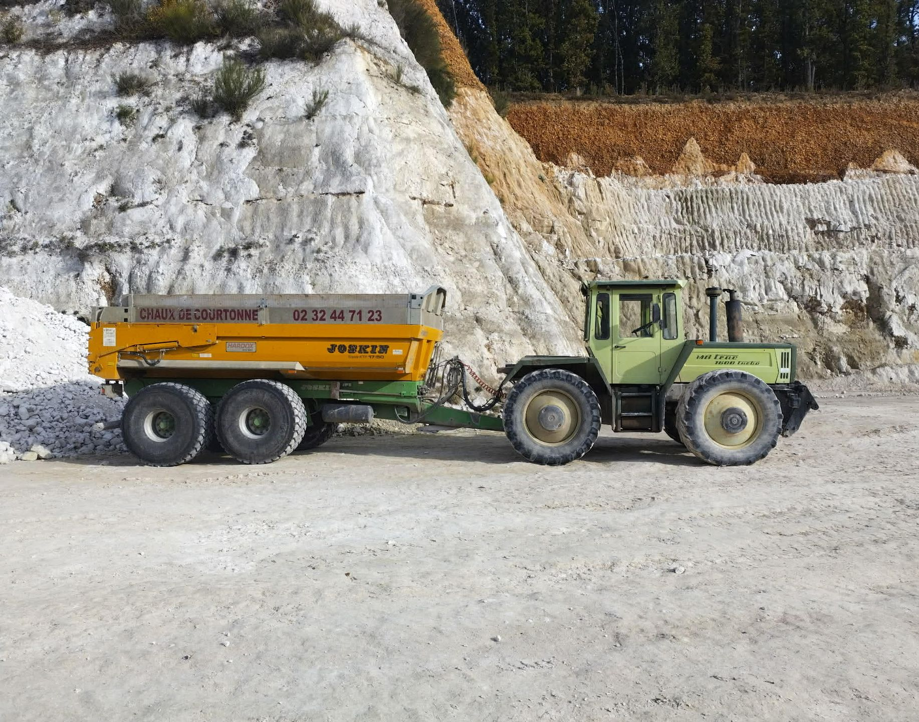 Un tracteur vert clair tirant une remorque benne jaune au pied d'une carrière de craie blanche.