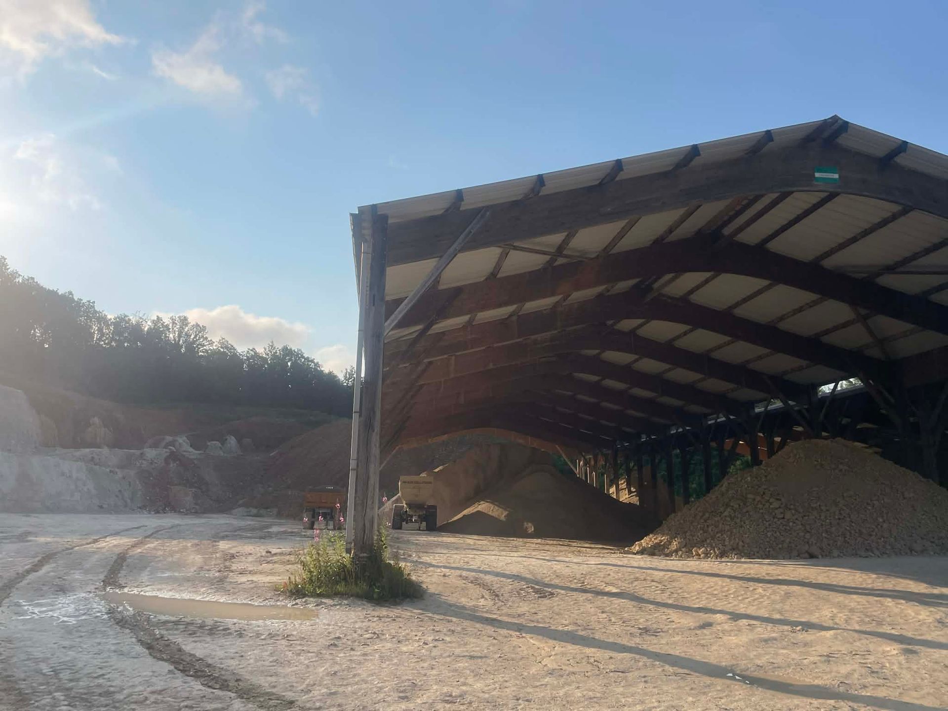 Un hangar industriel ouvert sur les côtés, situé dans une gravière, abritant des tas de sable et de pierres sous un grand toit.