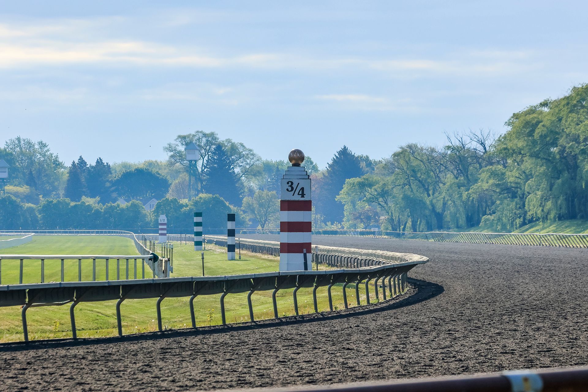 Une piste de courses hippiques en terre battue longe une barrière verte, avec un poteau rayé rouge et blanc marqué « 3/4 » au centre.