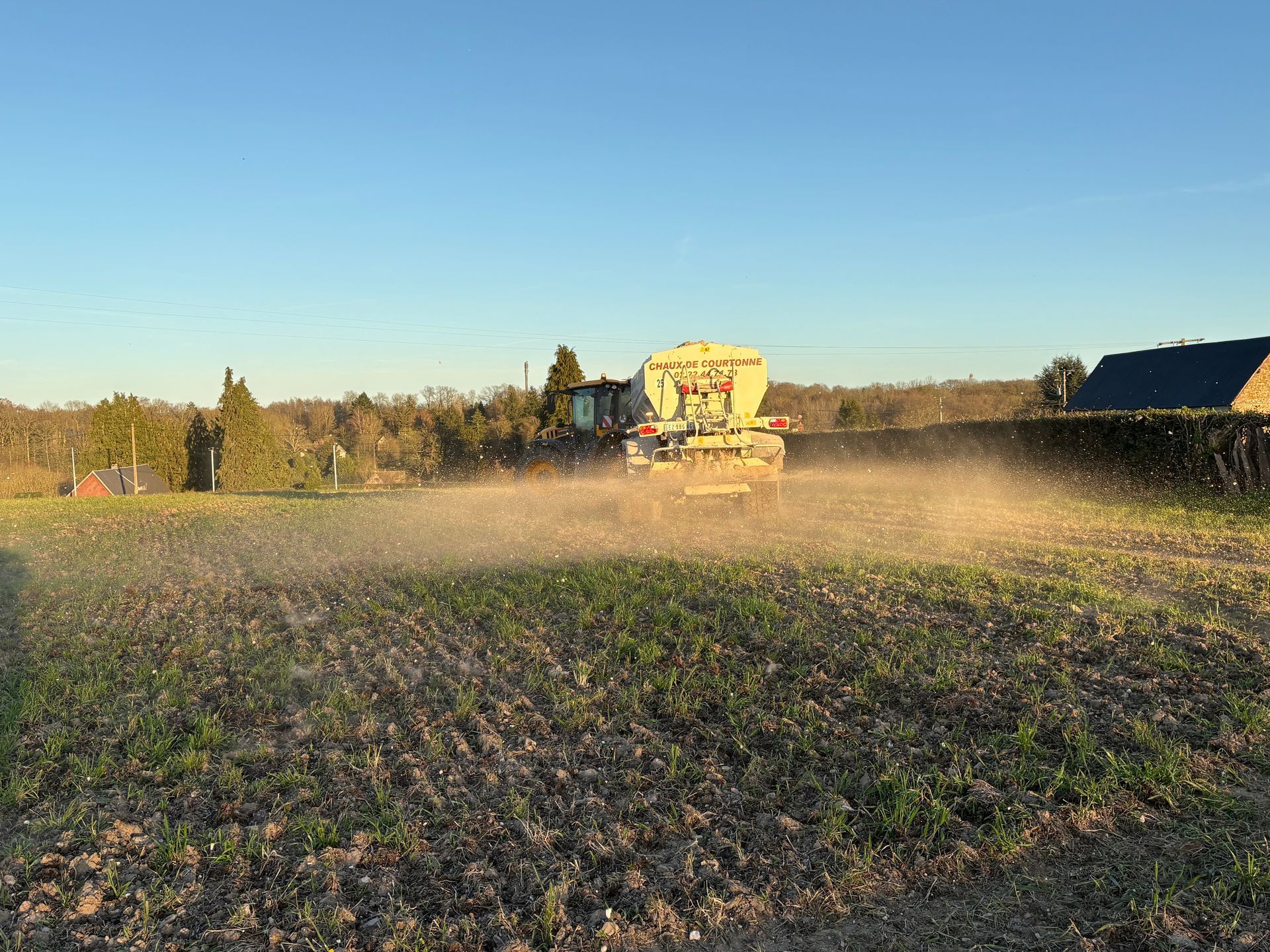 Un tracteur traverse un champ, répandant de la chaux sous un ciel bleu azur.