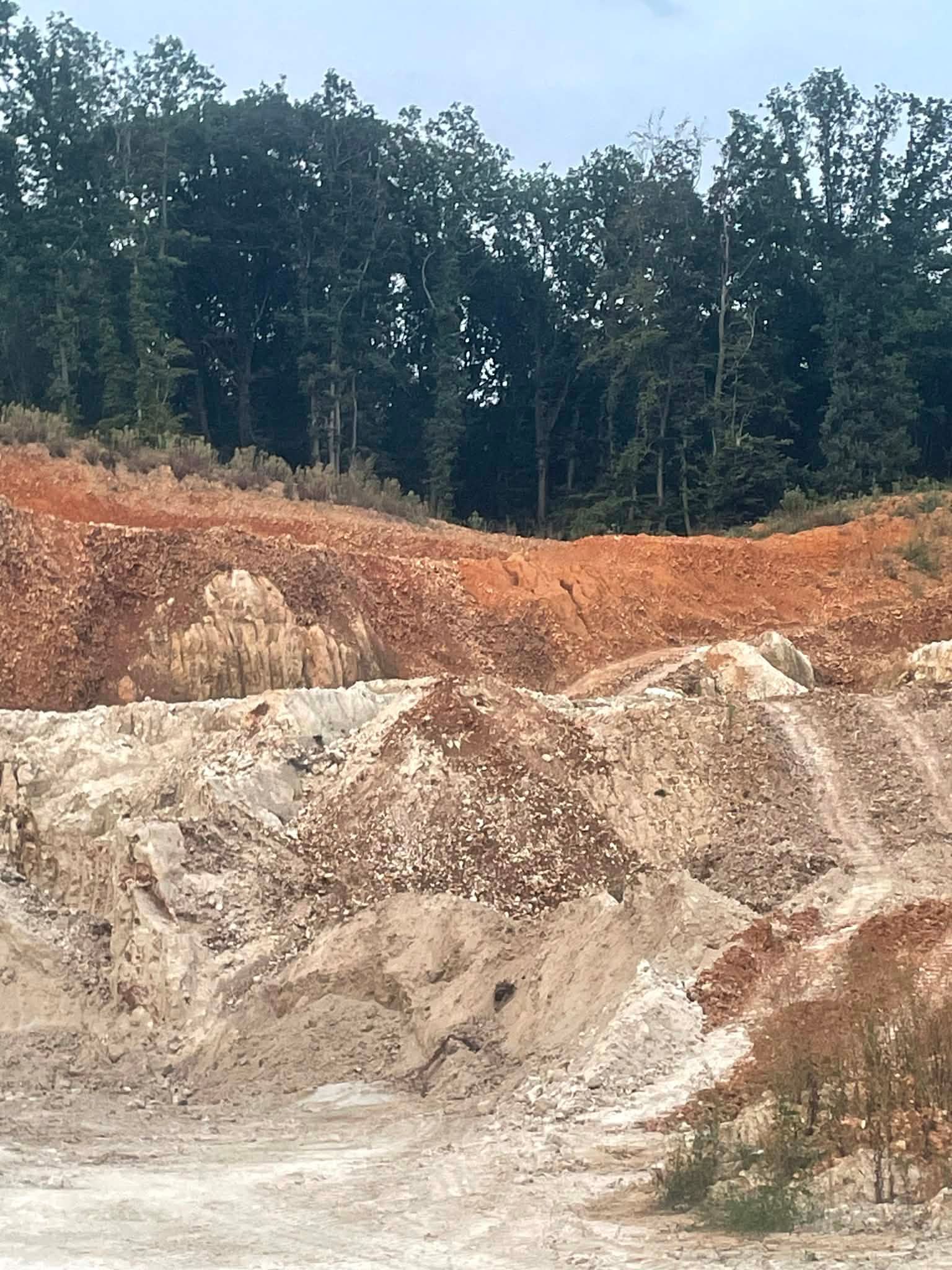 Une carrière révélant des couches de terre argileuse rouge sur un sol rocailleux clair, le tout adossé à une forêt dense et verdoyante.