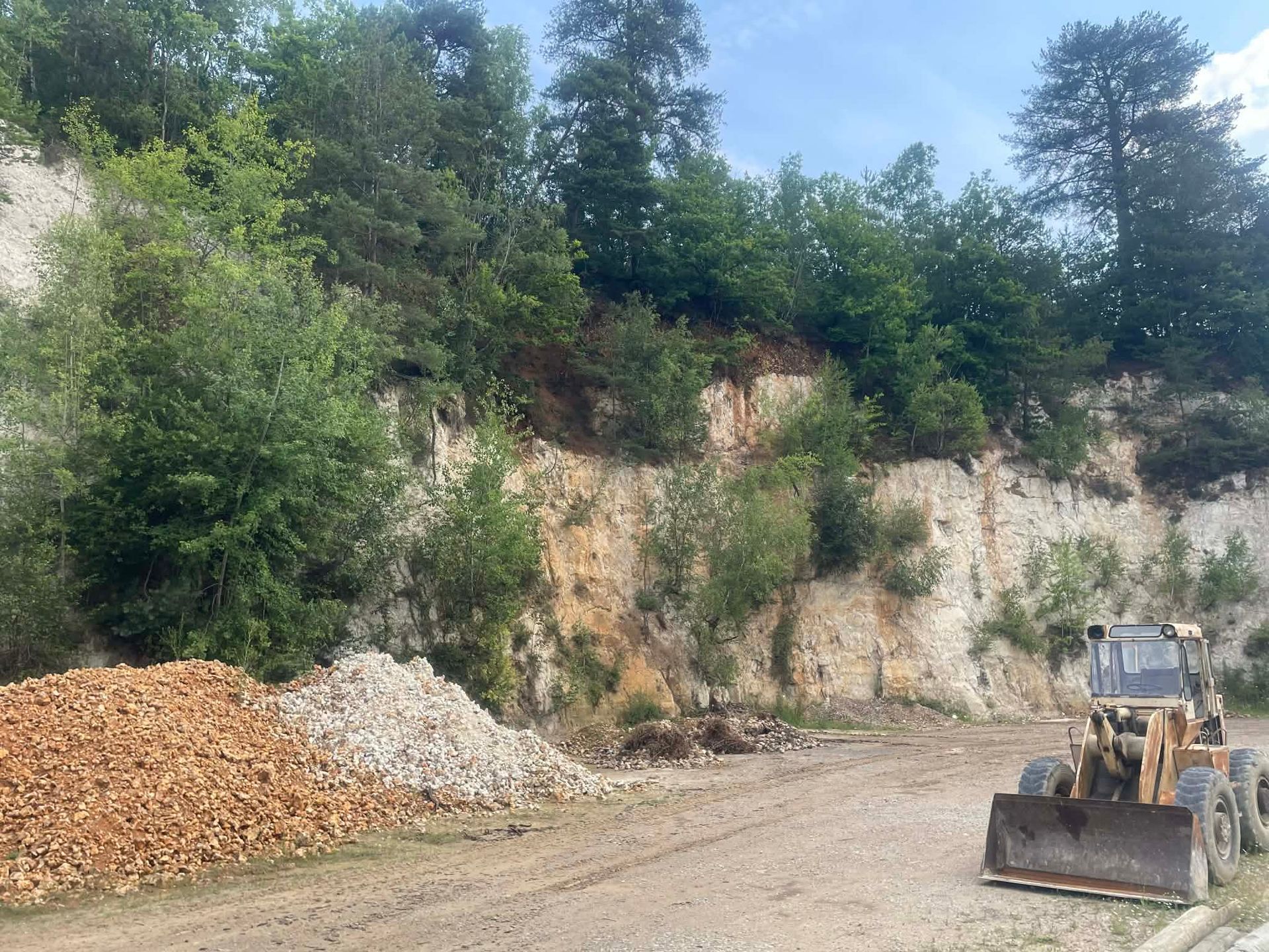 Un engin de chantier stationné devant une paroi rocheuse blanche d'une carrière, surmontée d'arbres, avec des tas de pierres au premier plan.