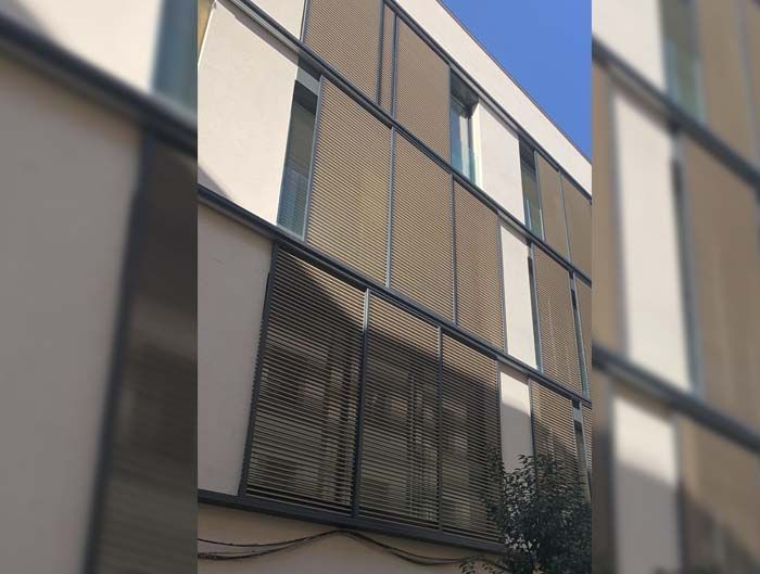 Fachada de edificio moderno con ventanas verticales y celosías oscuras contra un cielo azul.