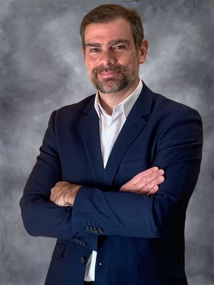Retrato de estudio de un hombre con traje azul marino y brazos cruzados, de pie sobre un fondo gris.