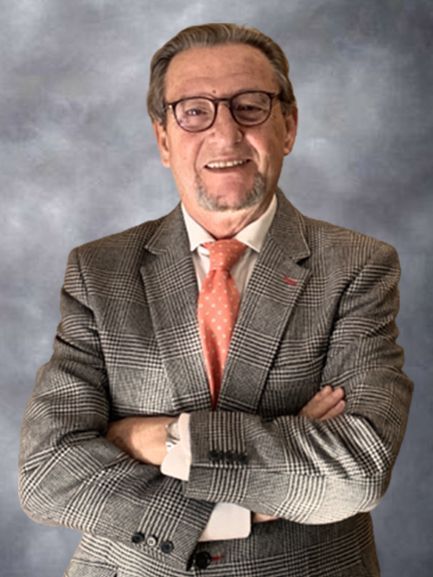 Hombre sonriente con gafas y traje gris a cuadros con corbata color coral, de pie con los brazos cruzados sobre un fondo gris.