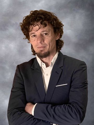 Hombre con traje oscuro y brazos cruzados, de pie frente a un fondo de estudio gris.
