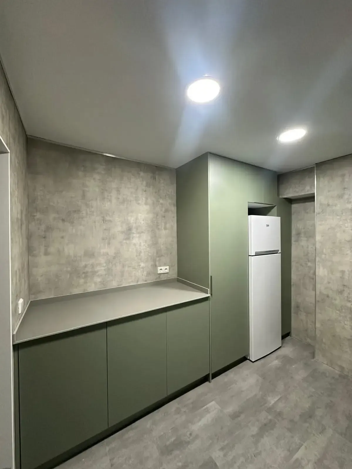 Cocina moderna con armarios verdes, paredes con aspecto de hormigón, frigorífico blanco y luces empotradas en el techo.