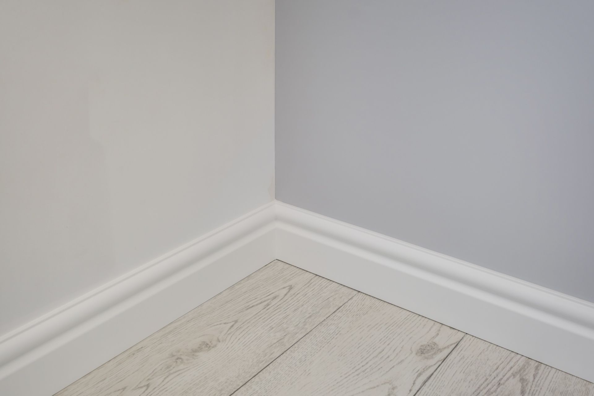 Esquina de una habitación con paredes grises, rodapié blanco y suelo imitación madera clara.