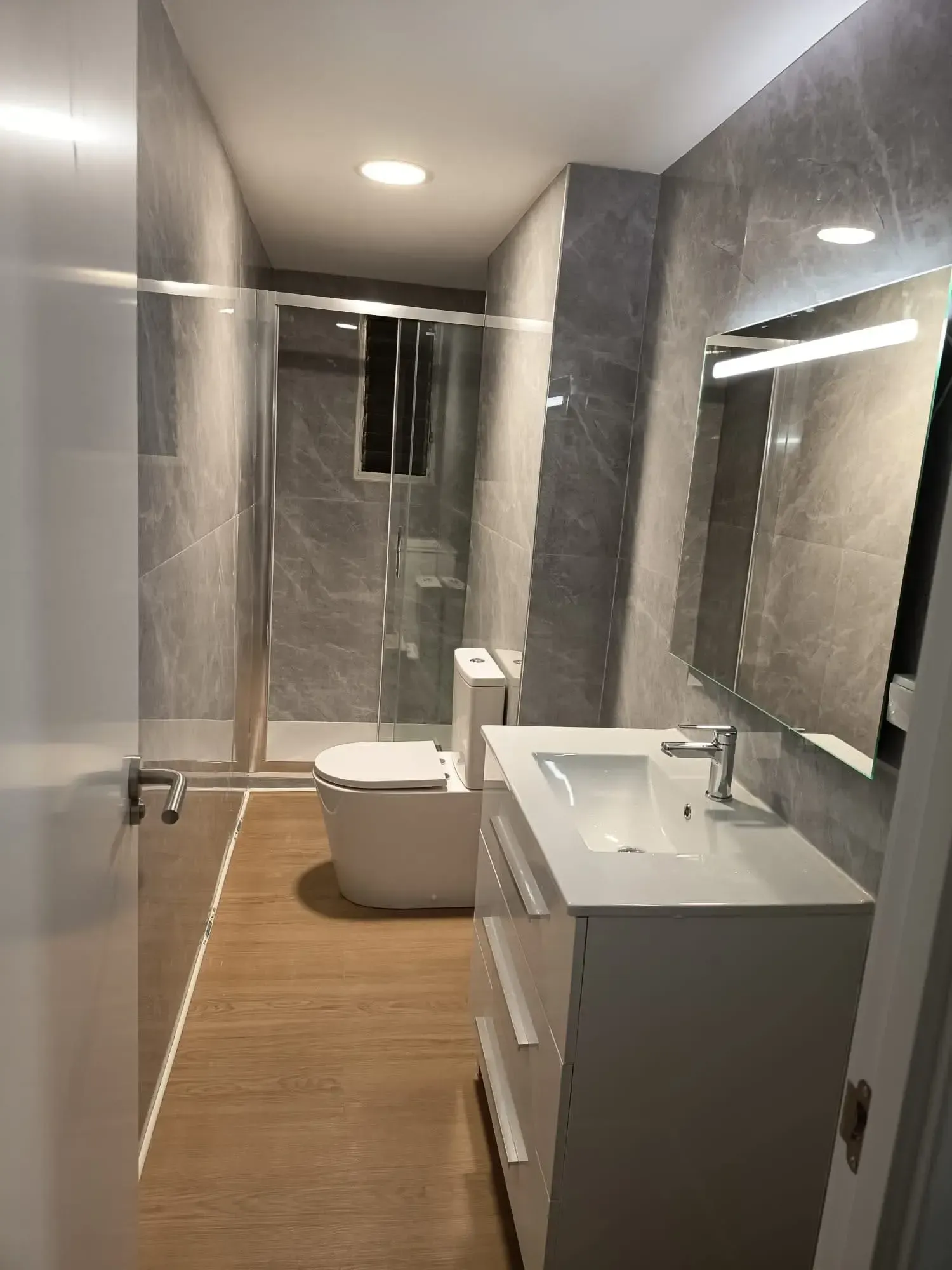 Baño moderno con ducha de cristal, bañera independiente y paredes de azulejos grises sobre suelo de madera.