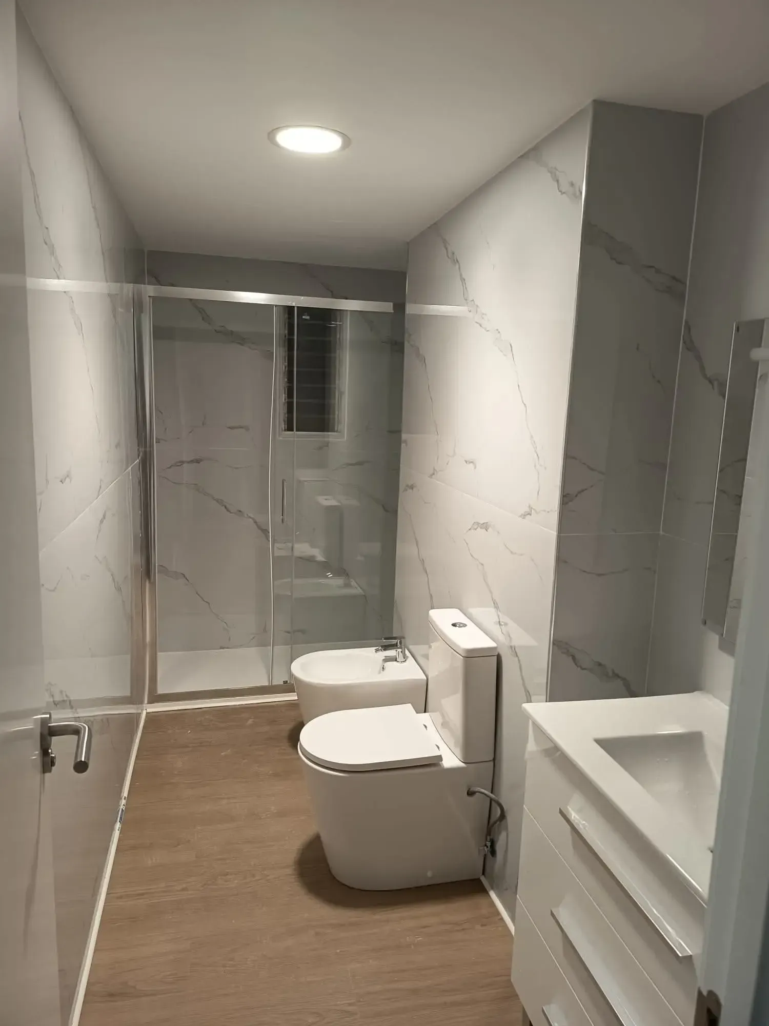 Baño moderno con ducha de cristal, inodoro y bidé blancos, paredes grises y suelo de madera.