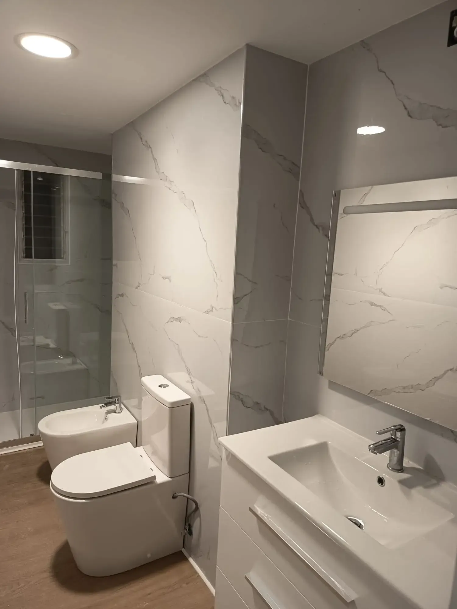 Baño moderno con inodoro, lavabo con mueble, espejo grande y paredes revestidas con azulejos que imitan el mármol.