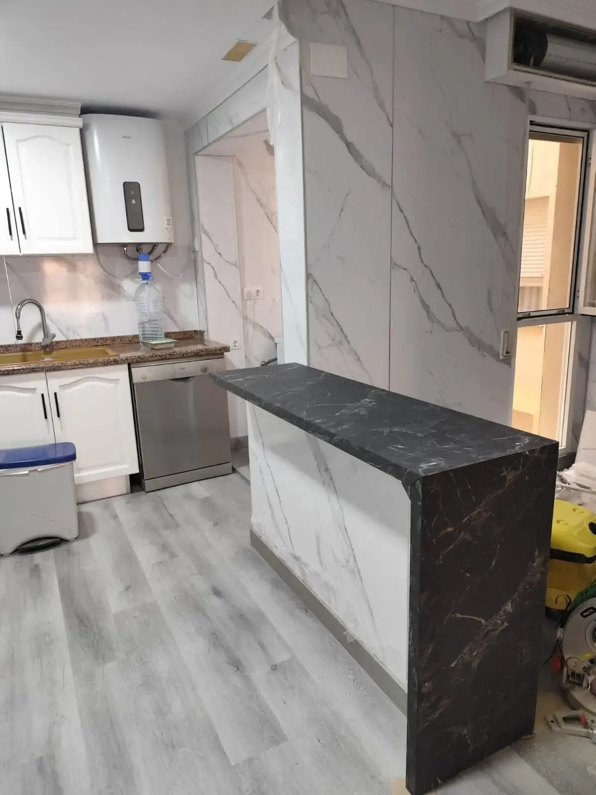 Cocina moderna en proceso de renovación con gabinetes blancos, piso de madera gris y una isla de cocina negra.