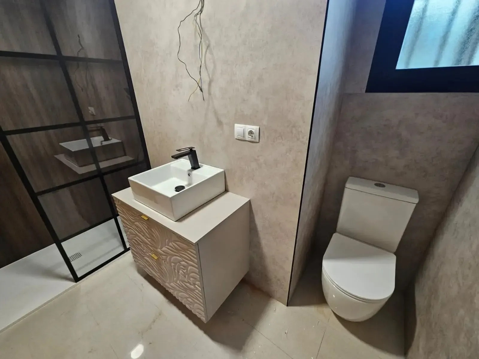 Baño pequeño con lavabo, inodoro, cabina de ducha y pared agrietada cerca de una ventana.