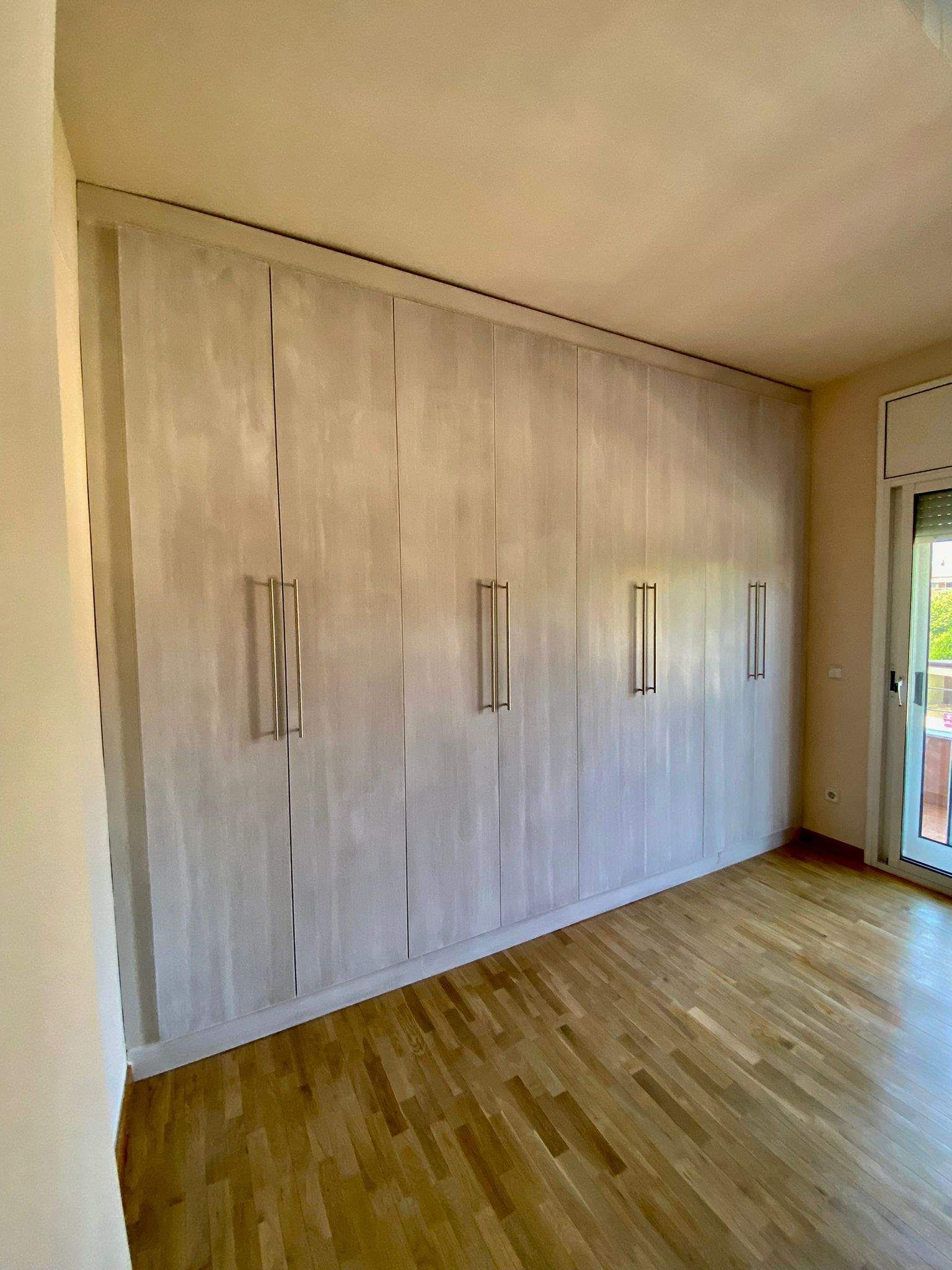 Armario empotrado gris claro con tiradores plateados contra la pared. Suelo de madera. La habitación tiene ventana.