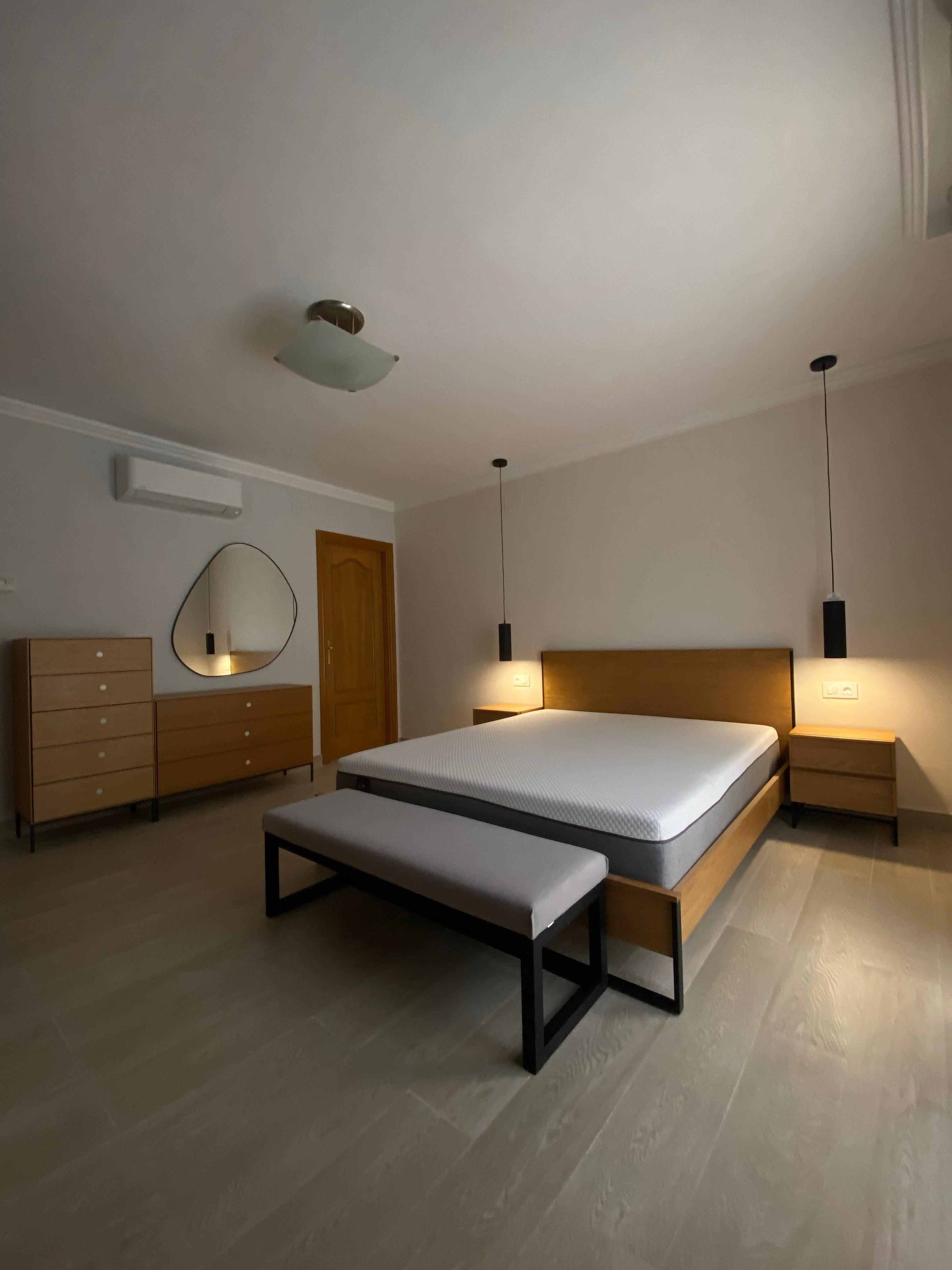 Dormitorio con muebles de madera: cama, cómoda, mesitas de noche y banco. Suelos y paredes de madera clara.