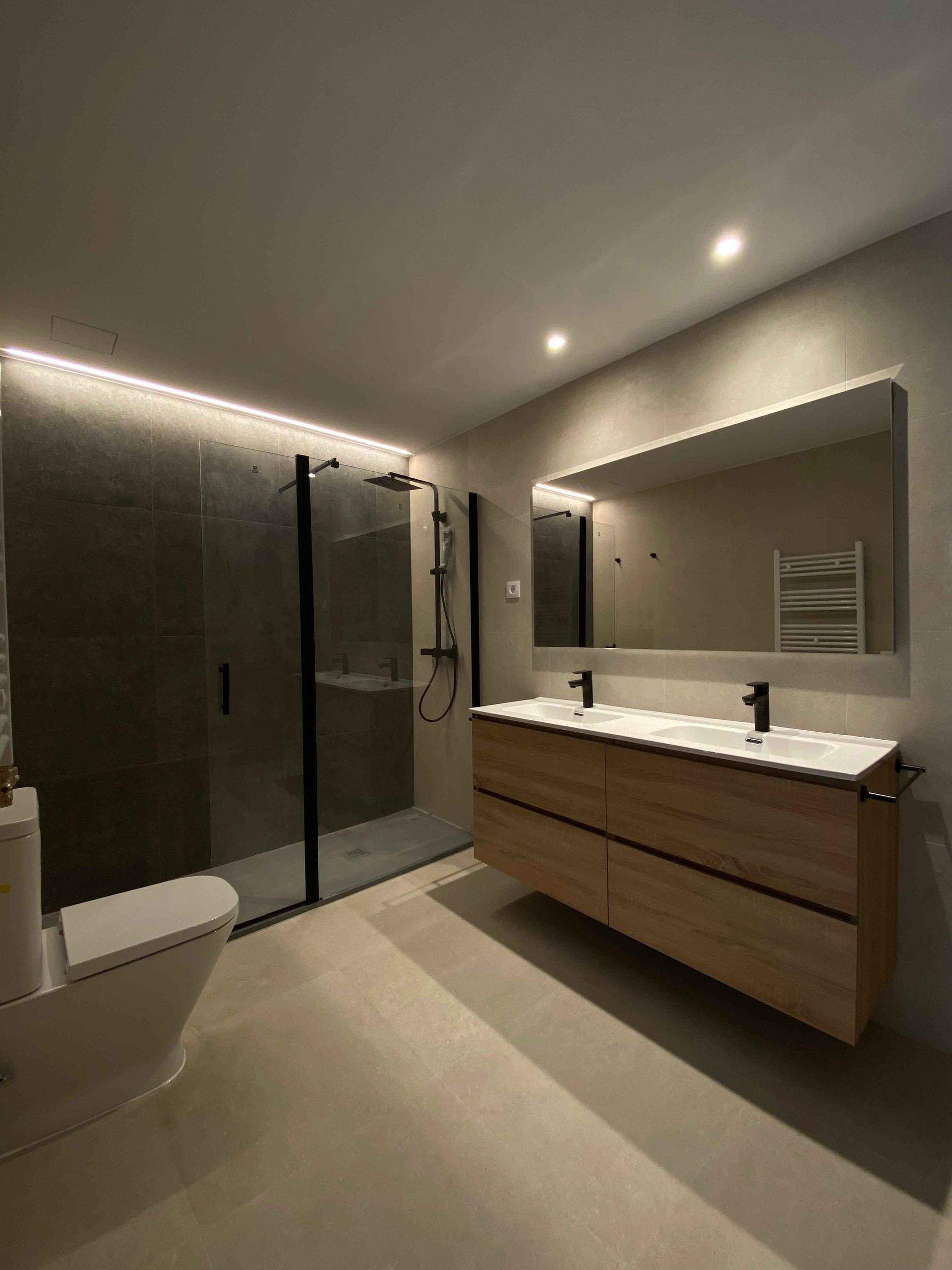 Baño moderno con ducha de cristal, lavabo doble e inodoro. Detalles en beige y negro.