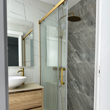 Interior del baño con accesorios dorados, puerta de ducha de vidrio y azulejos de mármol gris y blanco.