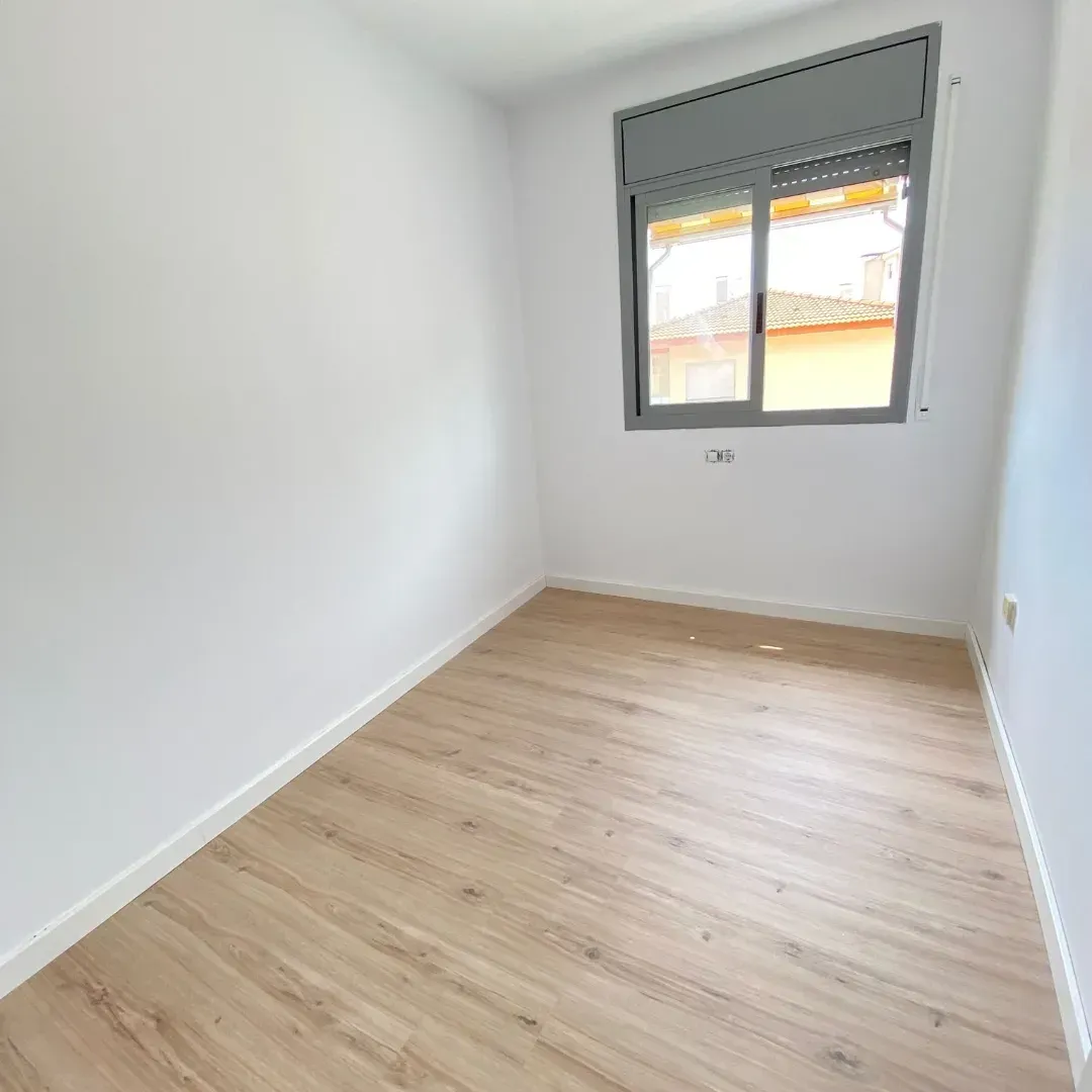 Habitación vacía con pisos de madera clara, paredes blancas y una ventana con persianas grises cerradas.