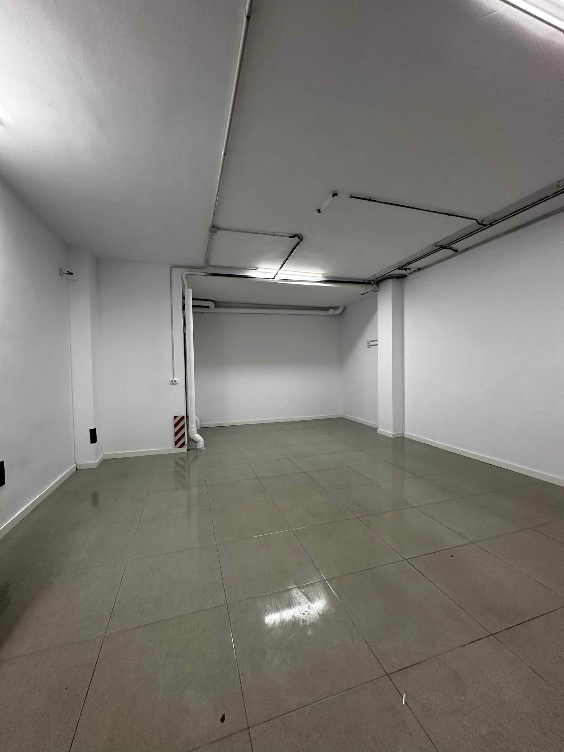 Habitación vacía, de paredes blancas, con suelo gris y tuberías del techo a la vista.