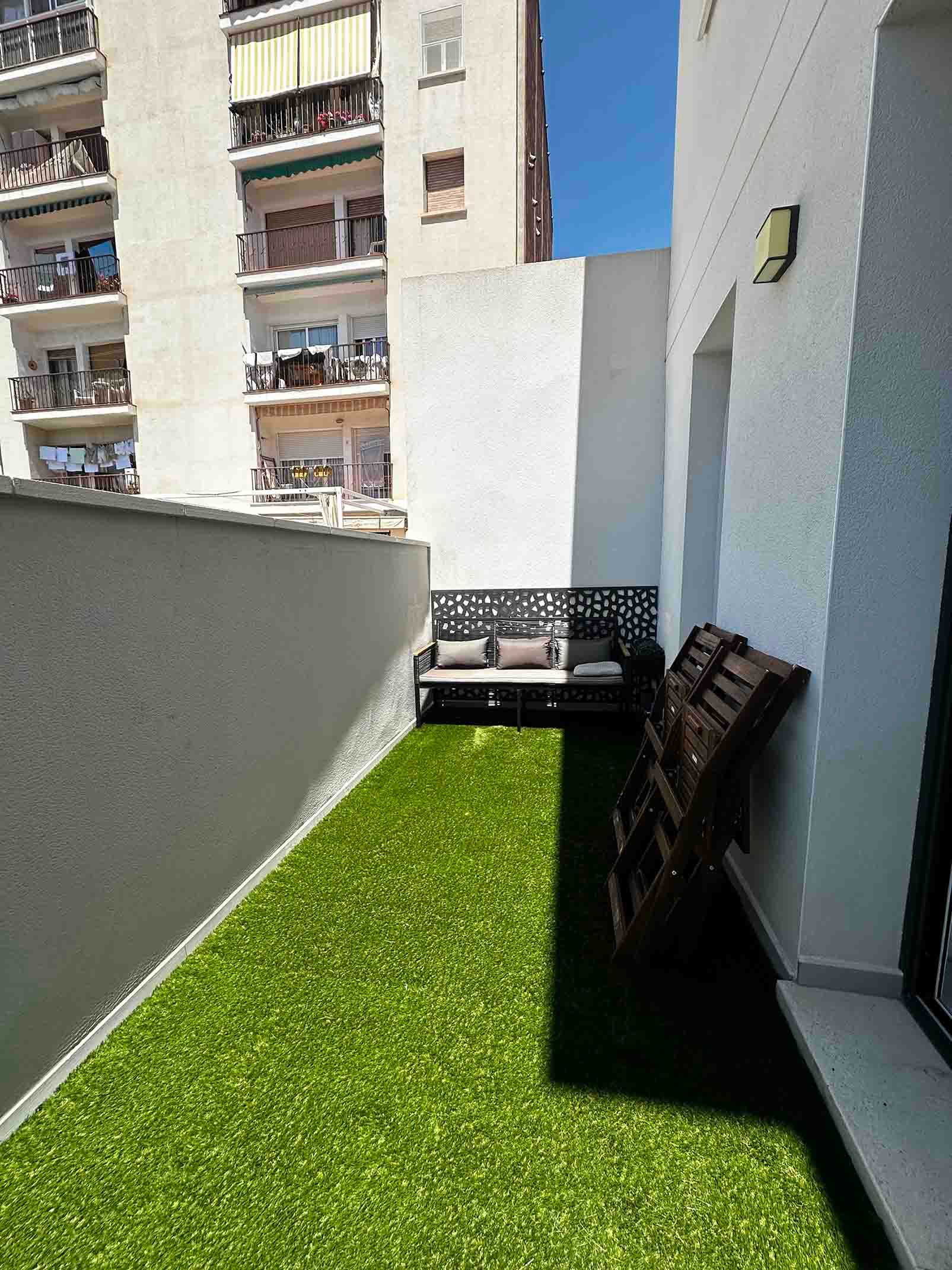 Pequeño espacio exterior con césped artificial, un banco y una silla plegable, con un edificio al fondo.