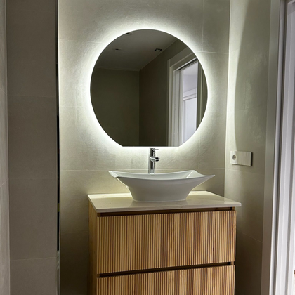 Baño con espejo redondo iluminado, lavabo sobre encimera y mueble de madera clara.