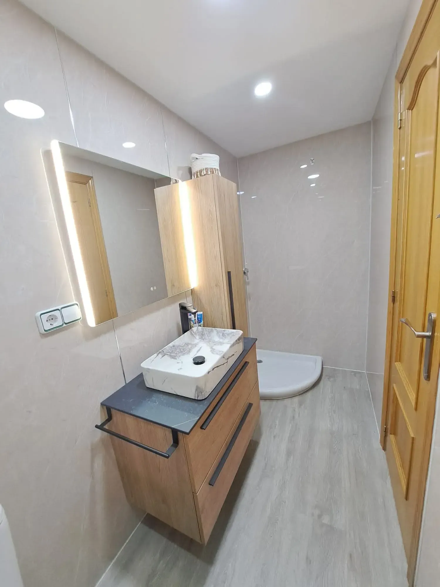 Baño moderno con tocador de madera, espejo de pared, lavabo, inodoro y puerta de ducha en tonos beige.