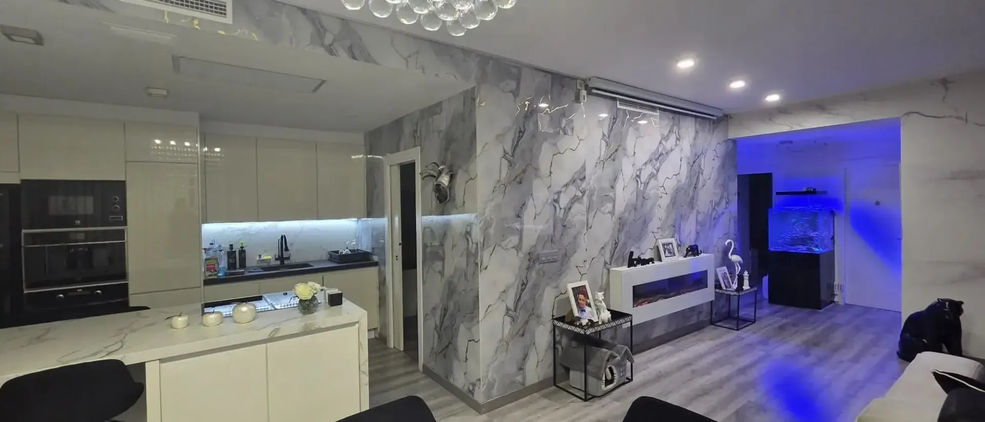 Cocina y sala de estar modernas con paredes de mármol, gabinetes blancos e iluminación ambiental azul.