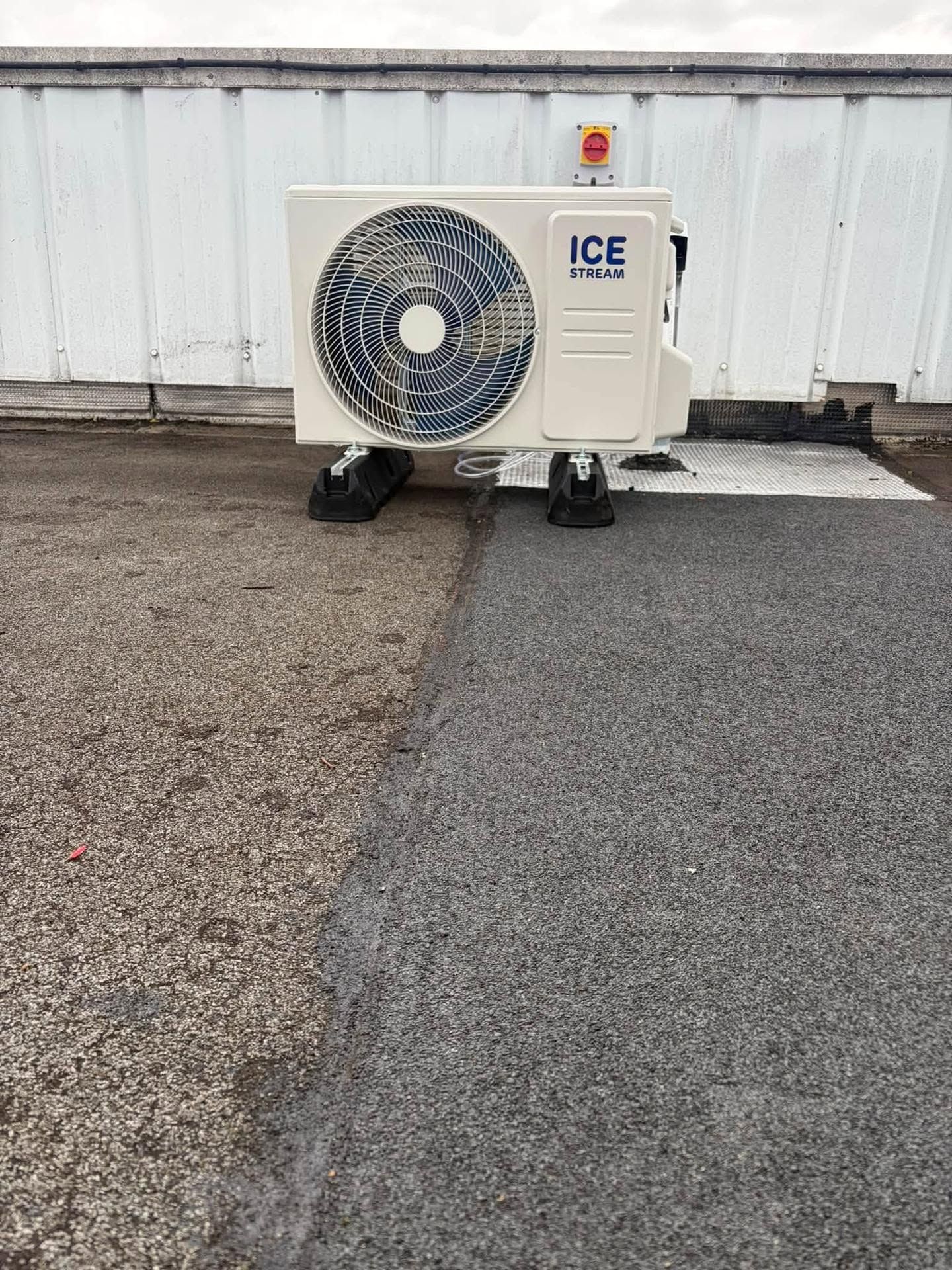 Unité de climatisation portable blanche sur roulettes noires contre un mur blanc, à côté de gravier et de pavés.