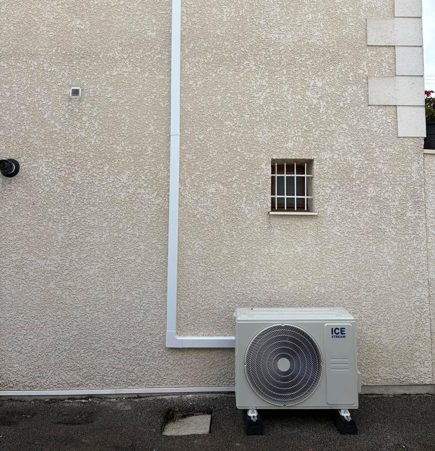 Mur extérieur avec tuyauterie blanche et petit climatiseur sous une fenêtre grillagée.