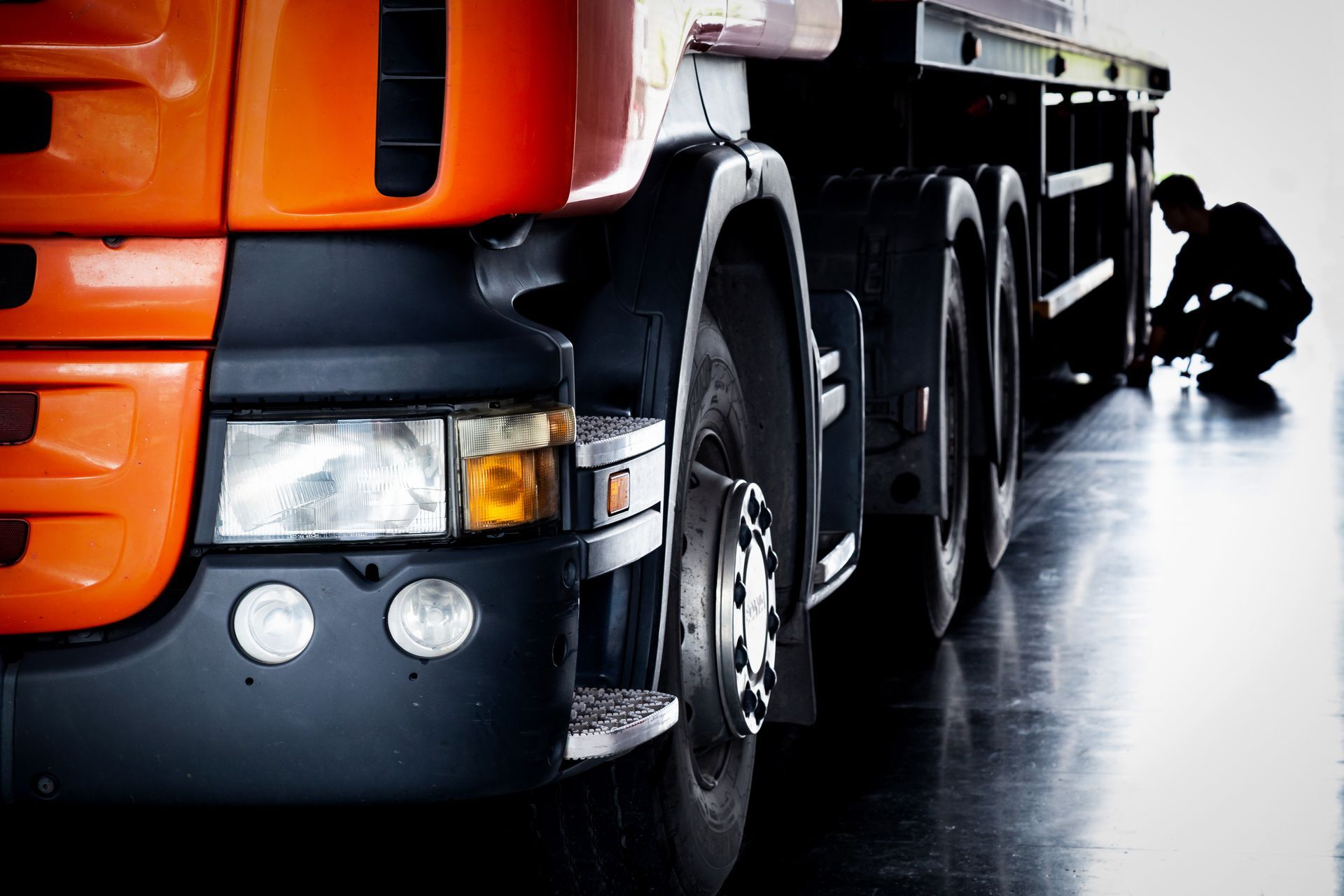Un camion orange et noir est stationné sur la chaussée mouillée, un ouvrier est accroupi près des roues arrière.