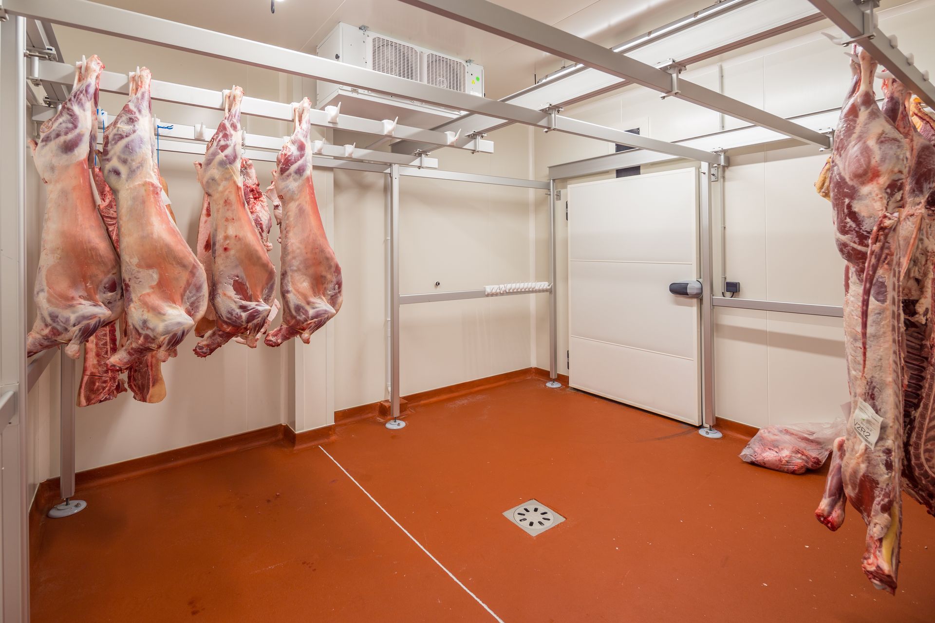 Chambre froide vide avec carcasses suspendues, sol orange, murs blancs et barres de plafond.