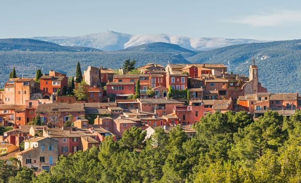 Vue panoramique d'une ville historique à flanc de colline, avec ses toits de tuiles rouges, ses clochers et les montagnes en arrière-plan.