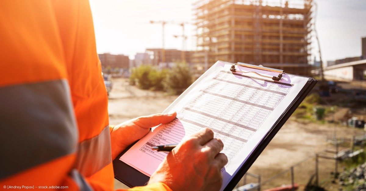 Ein Bauarbeiter in orangefarbener Sicherheitsweste schreibt auf einem Klemmbrett an einer Baustelle.