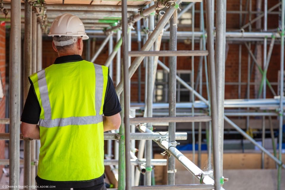 Ein Arbeiter in neongelber Sicherheitsweste und Schutzhelm steht auf einem Gerüst an einer Baustelle.