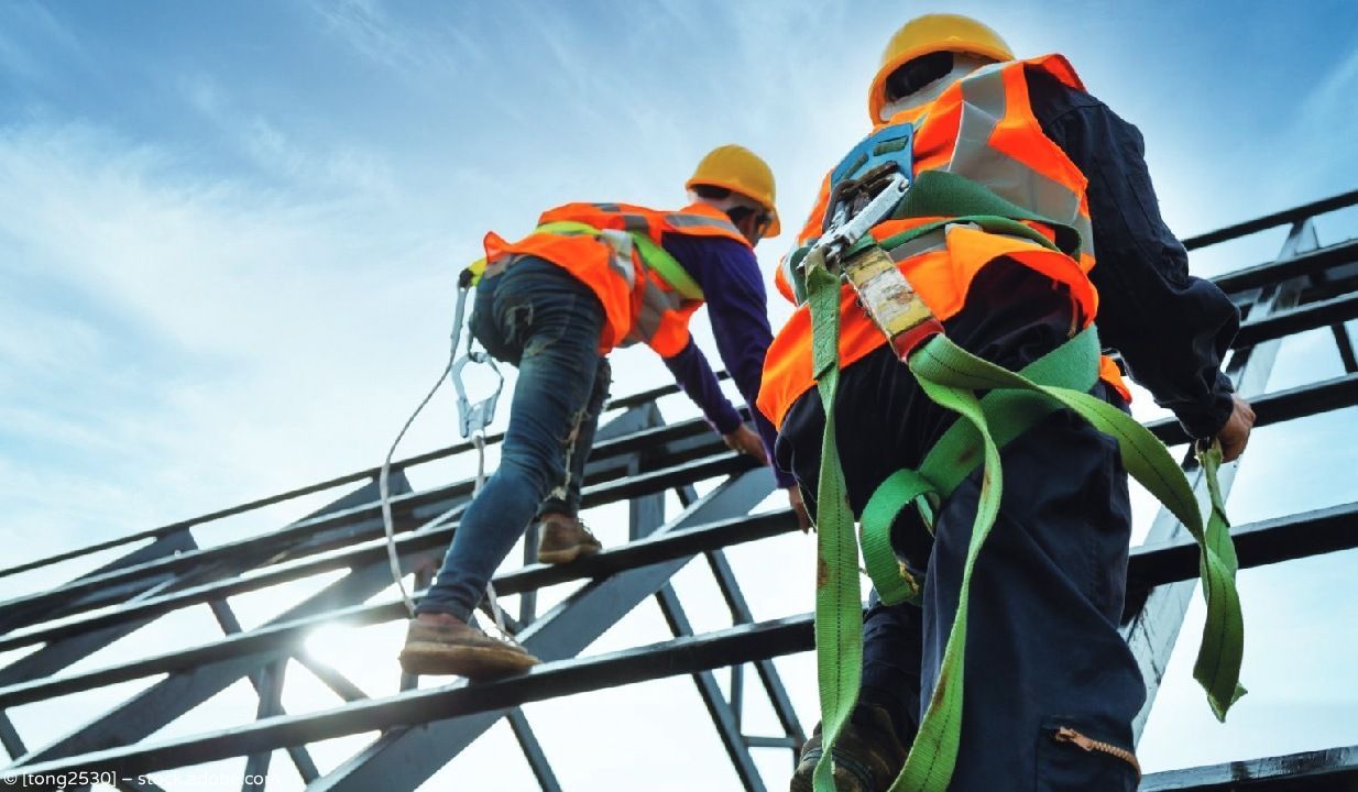 Arbeiter in orangefarbenen Sicherheitswesten und Schutzhelmen klettern auf einer Baustelle an einer Stahlkonstruktion hoch.