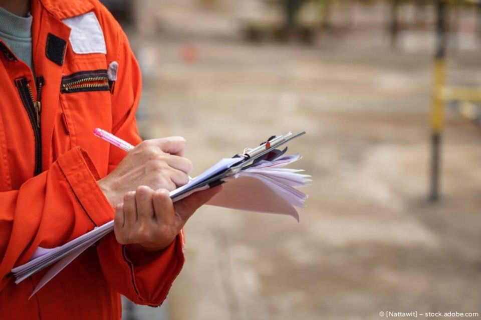 Ein Arbeiter in orangefarbener Sicherheitsjacke schreibt auf einem Klemmbrett an einer Baustelle.