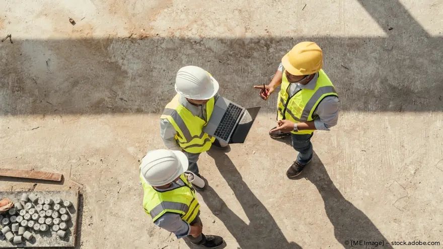 Drei Bauarbeiter mit Schutzhelmen und Sicherheitswesten prüfen Pläne auf einer Baustelle.