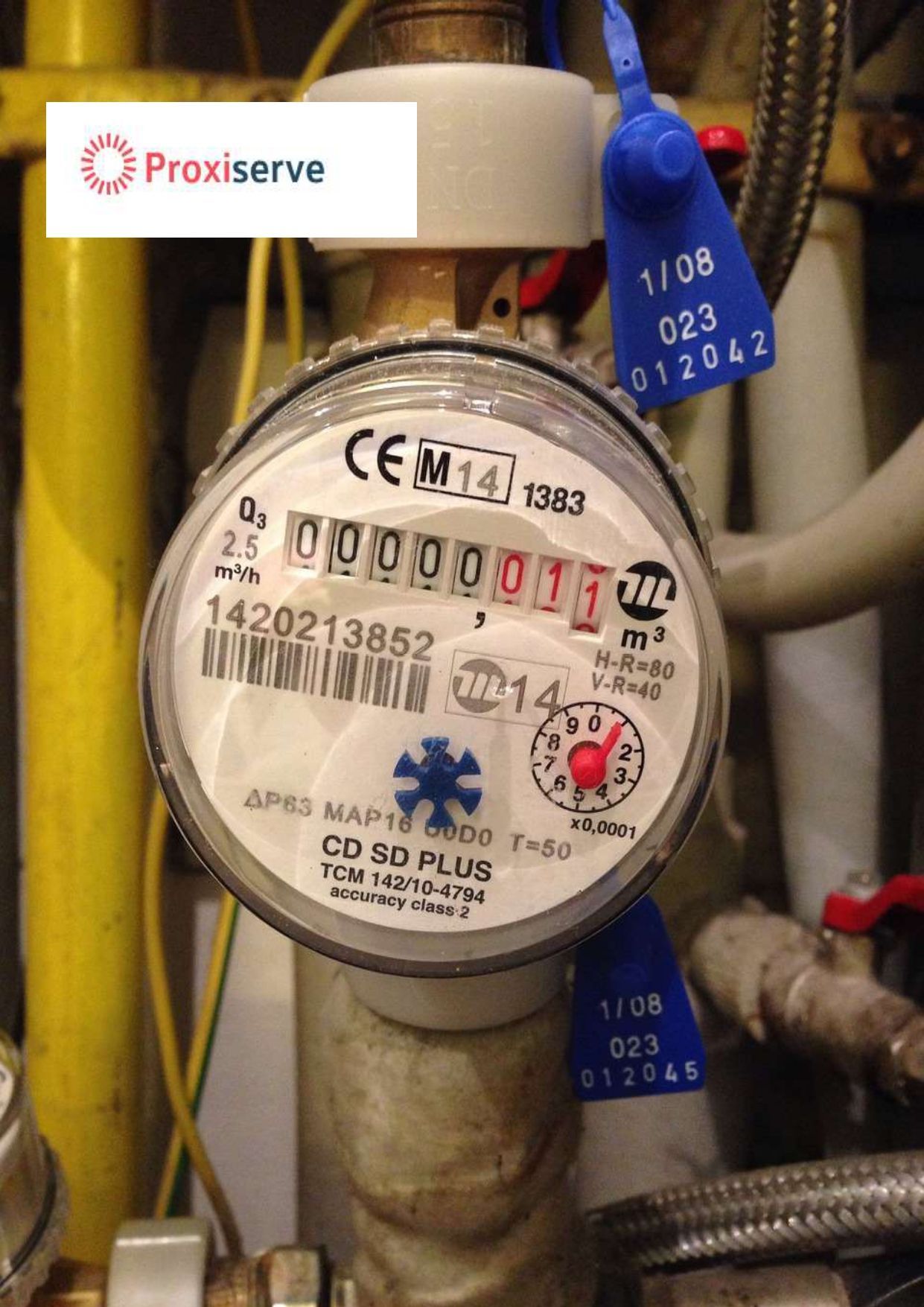 Gros plan sur un compteur d'eau à cadran blanc et rouge, parmi des tuyaux et des câbles jaunes.