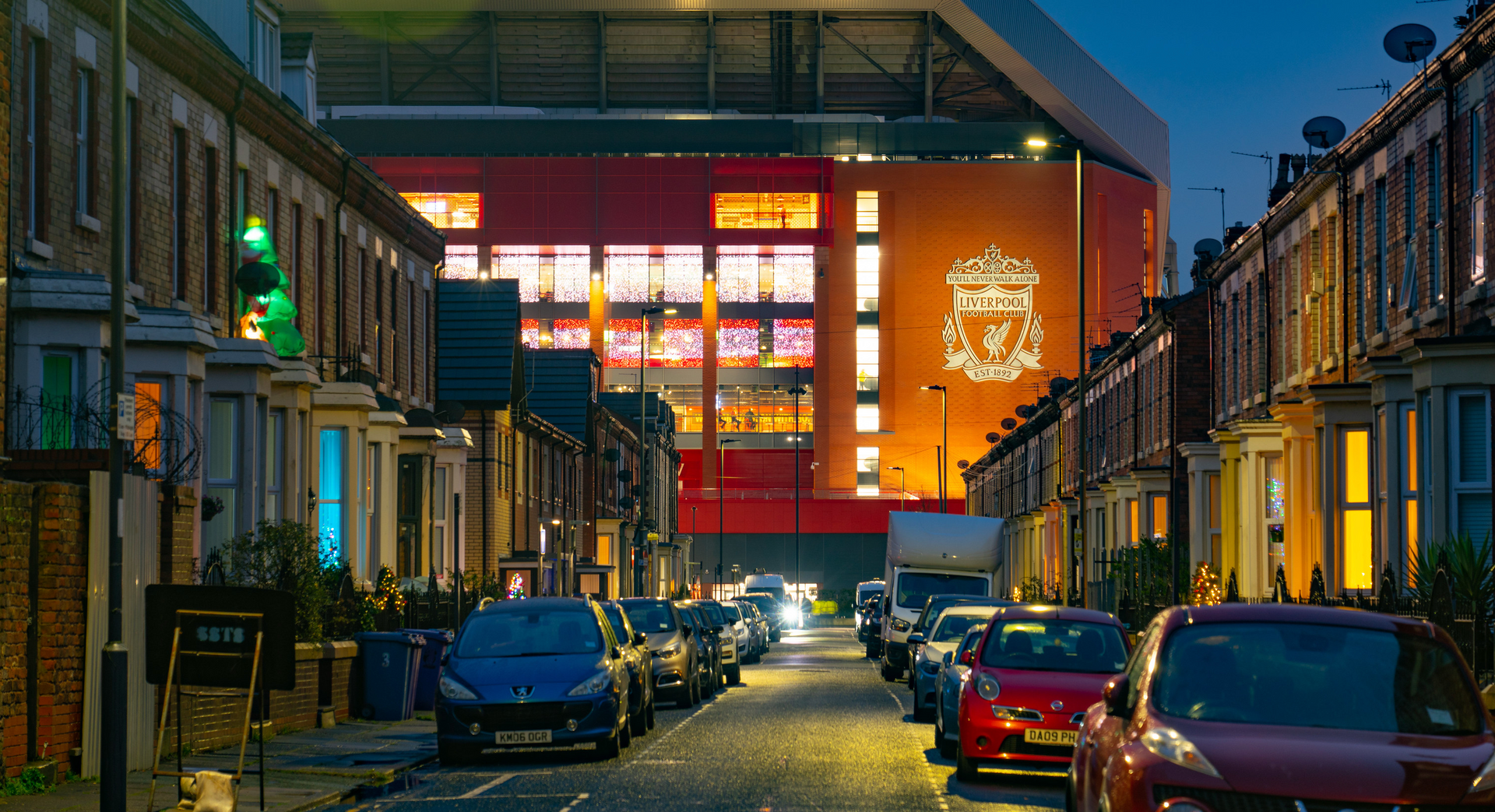 A row of cars are parked on Rockfield Road in front of Anfield stadium.