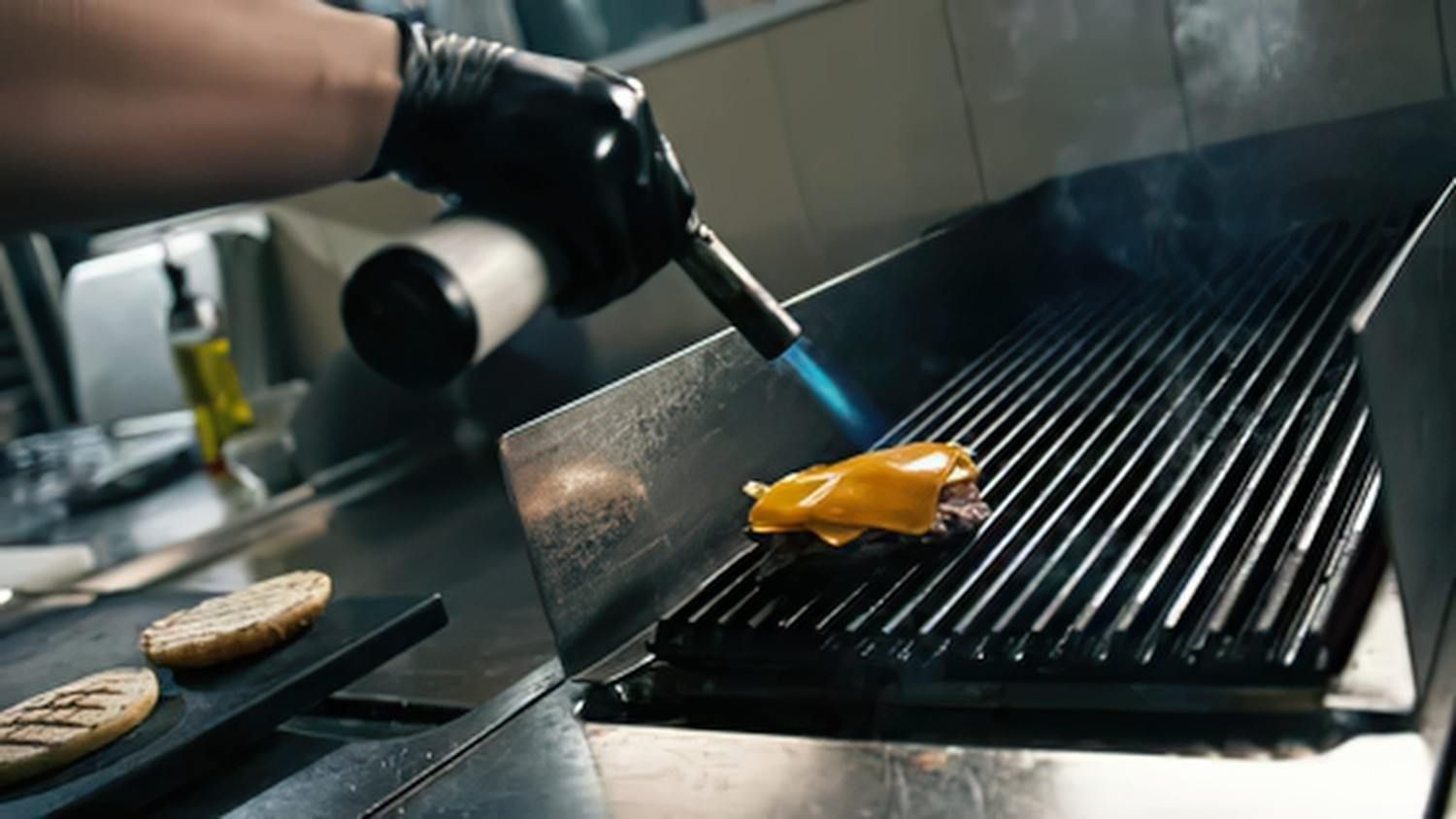 Un cocinero asa una hamburguesa y un pan en una parrilla humeante con unas pinzas