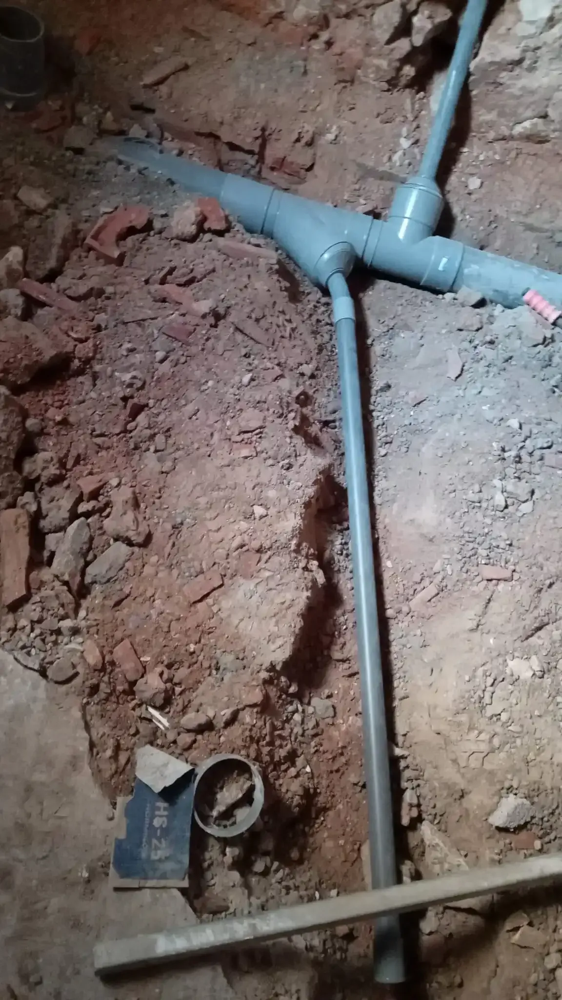 Zanja excavada con tuberías y accesorios de PVC gris en un muro de tierra, que muestra trabajos de fontanería en curso.