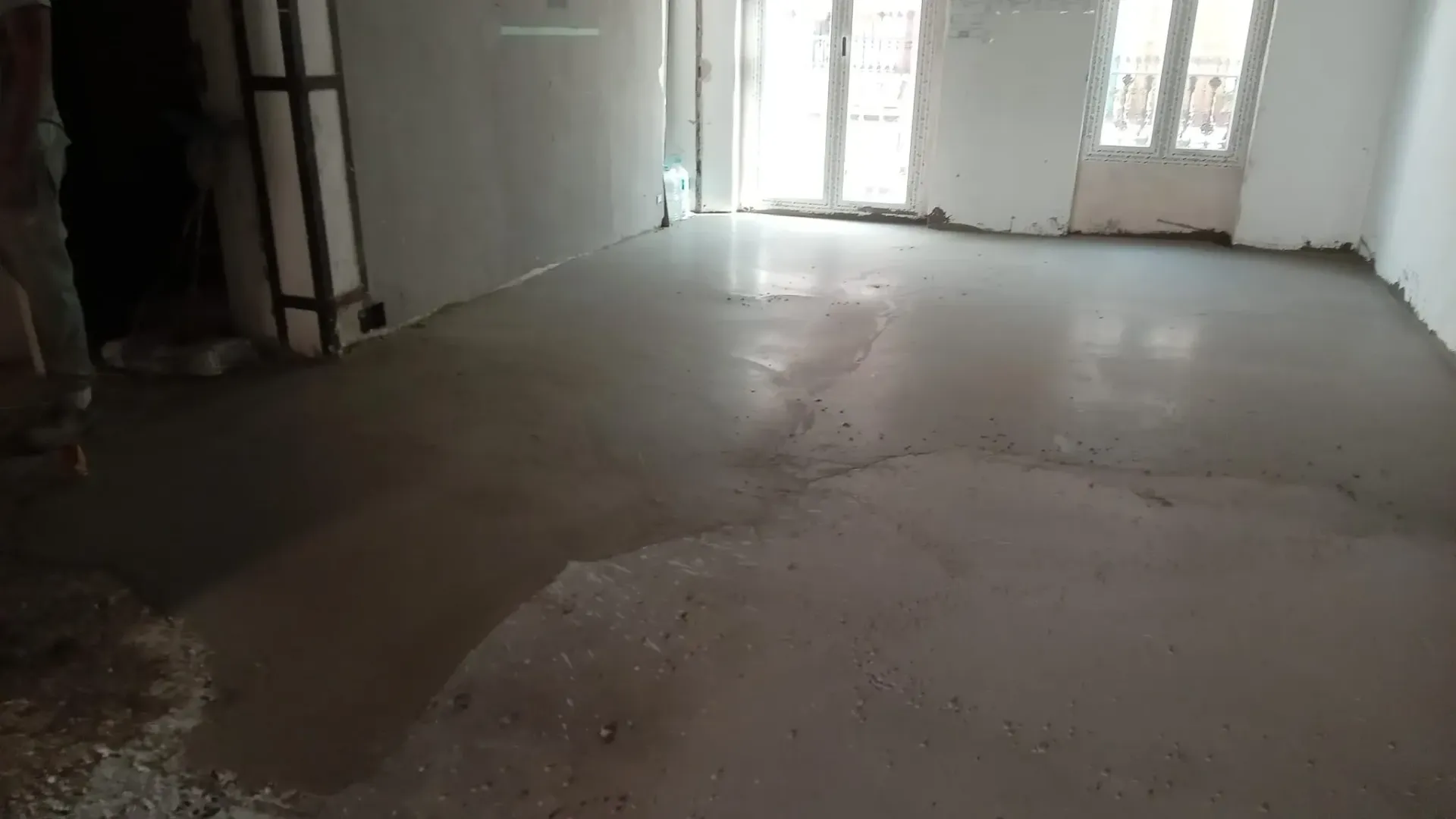 Habitación vacía sin terminar con suelo de hormigón visto y una puerta luminosa al fondo.