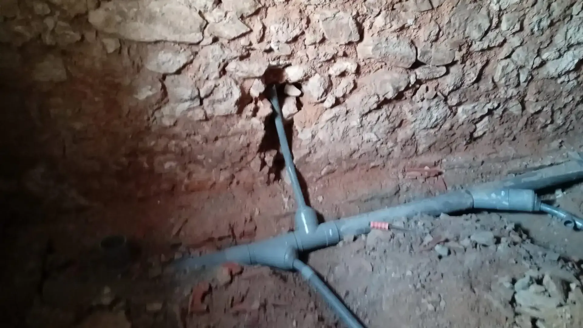 Tubería subterránea rota en una zanja de tierra junto a un muro de tierra irregular.