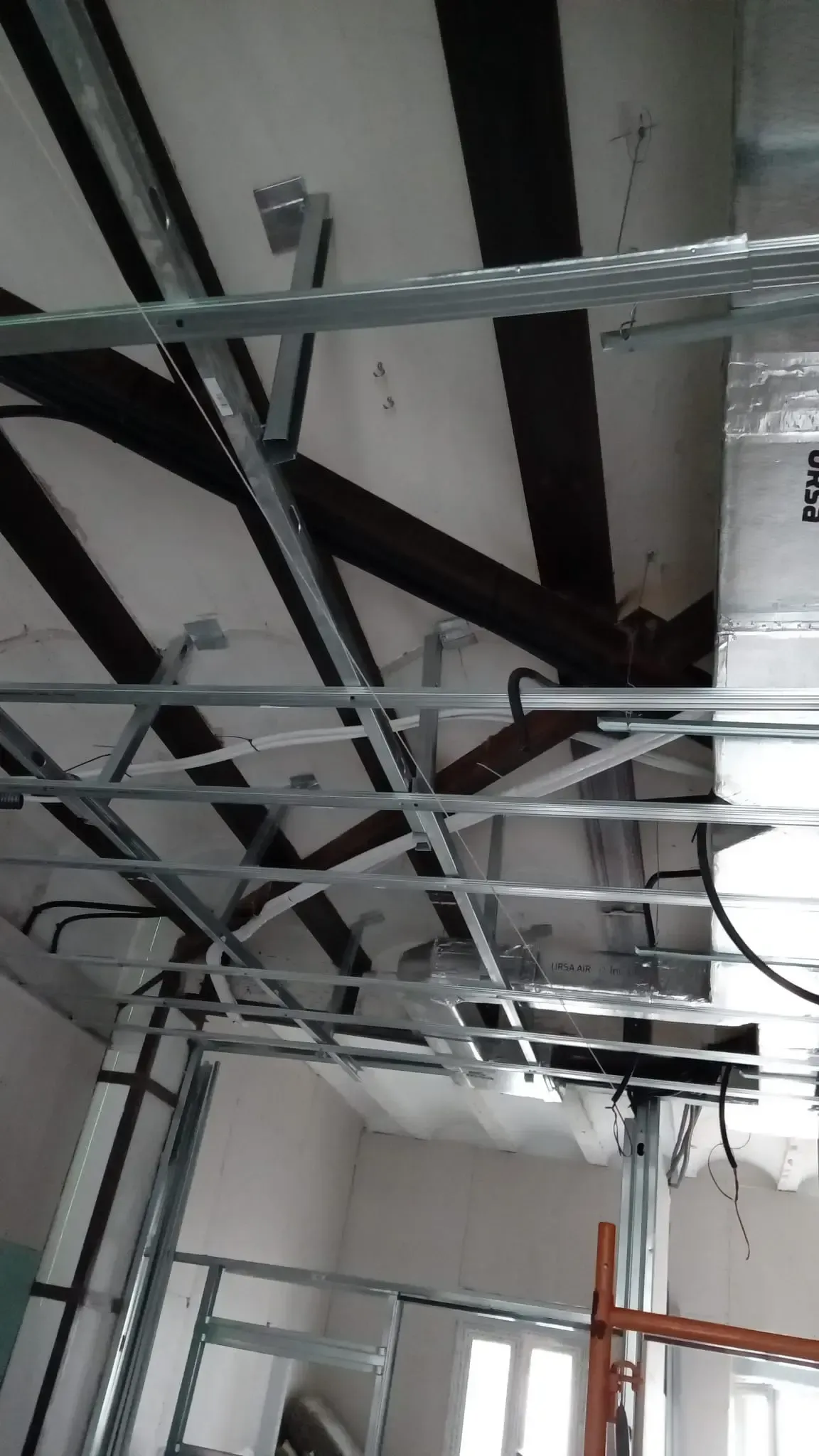 Techo a la vista con conductos metálicos, vigas negras y cables colgantes en una habitación interior sin terminar.