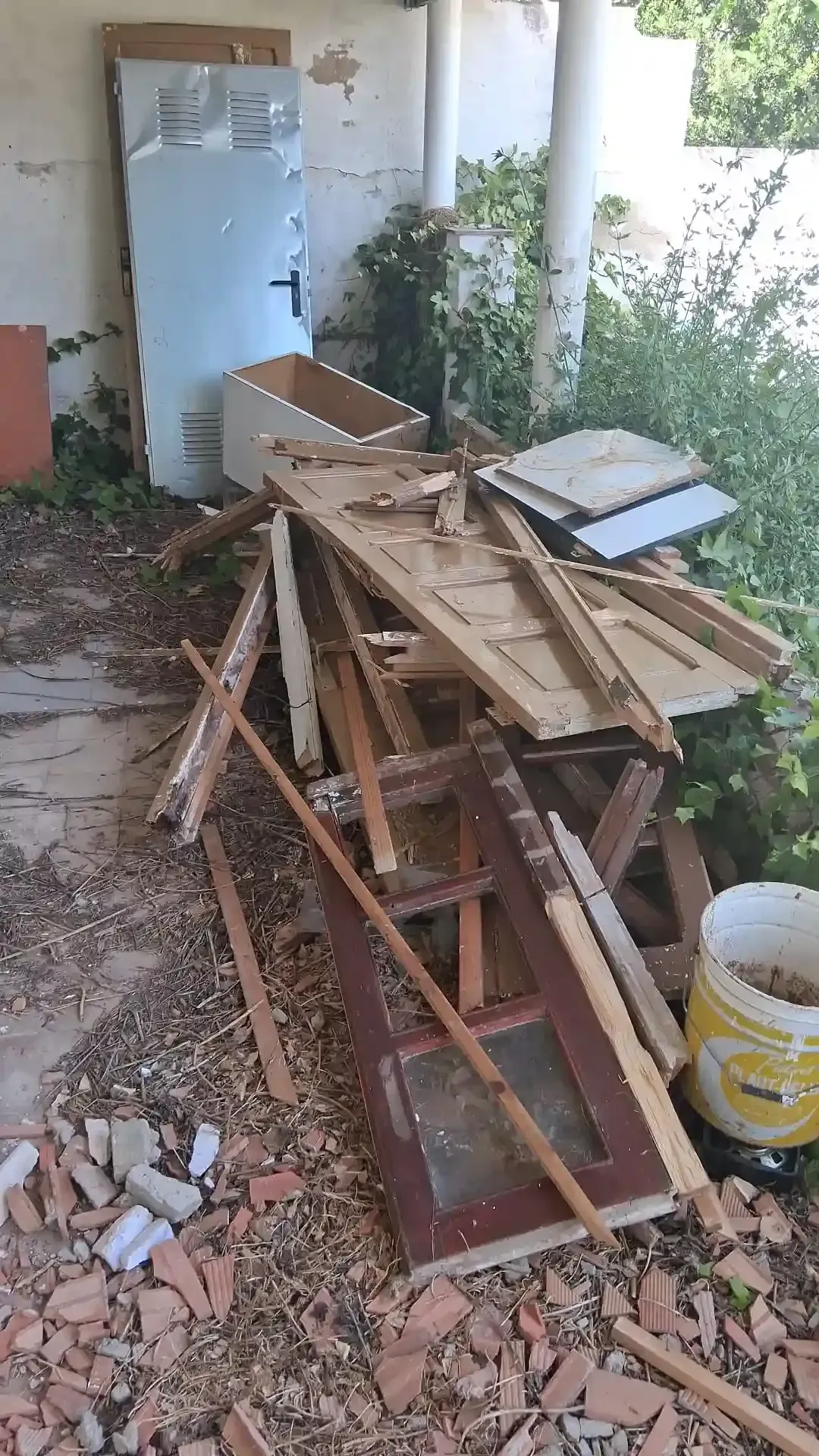 Montón de tablas de madera rotas y escombros en un pequeño patio exterior, junto a una puerta y una pared blancas.