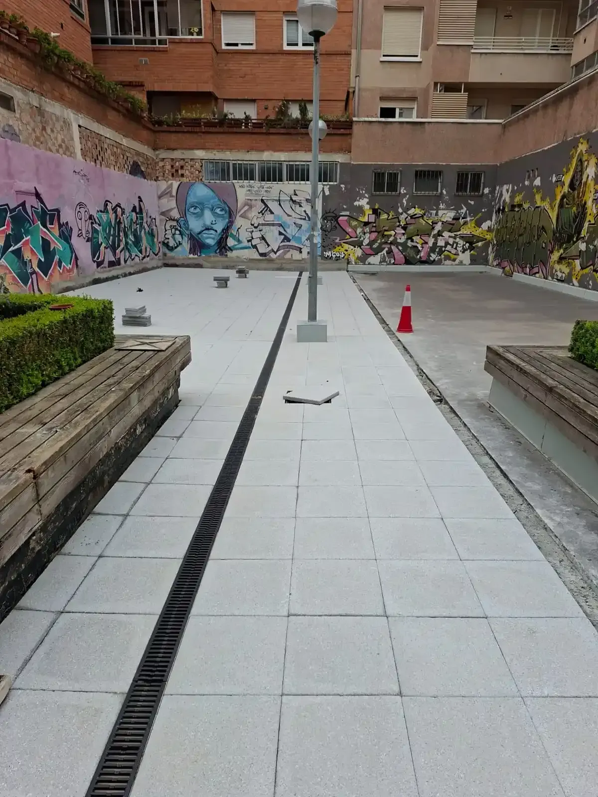 Patio urbano con camino de baldosas, jardineras de ladrillo, muro para grafitis y dos conos de tráfico.