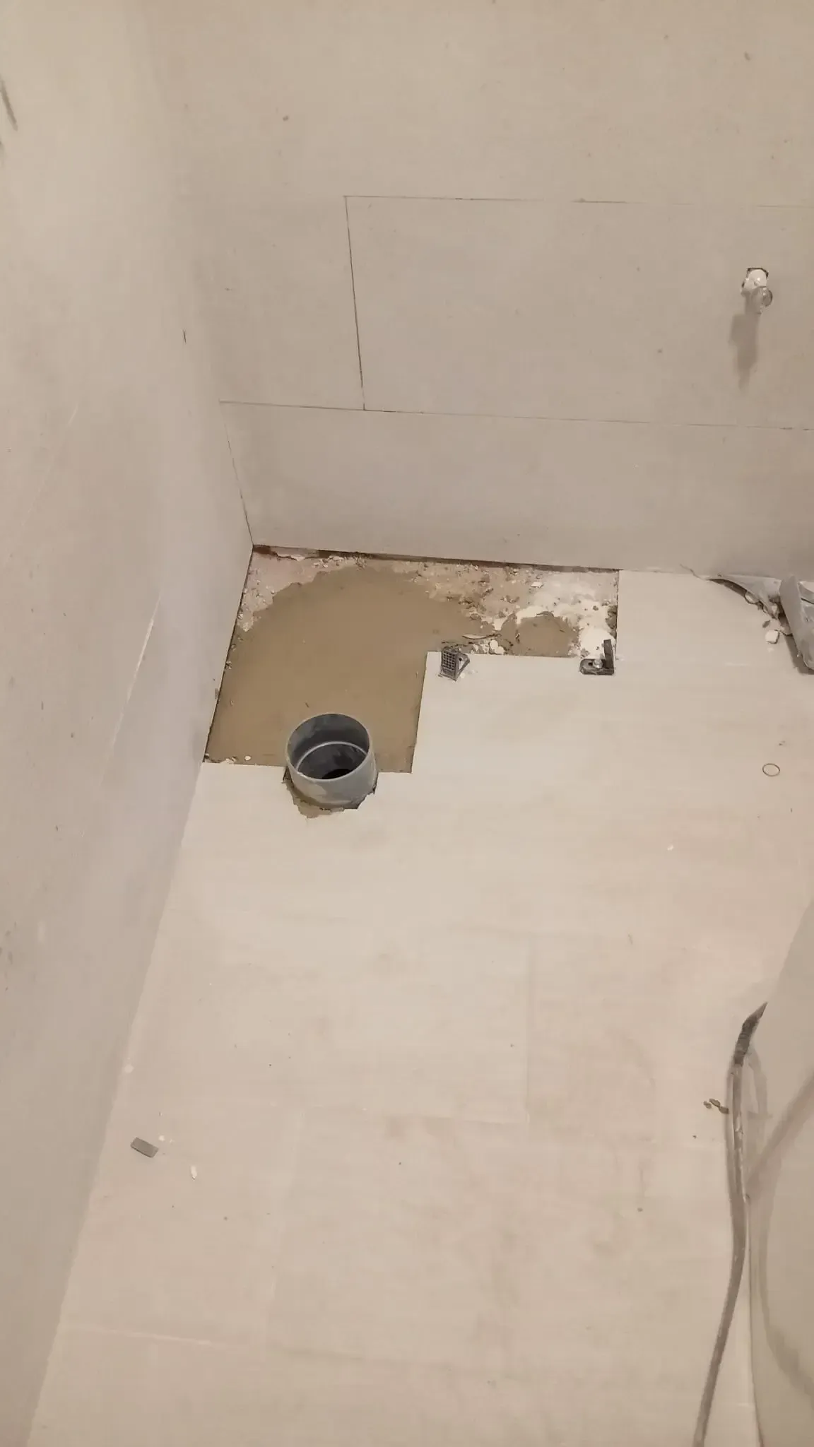 Pared y suelo blancos dañados con agujero y tubería expuestos en un pequeño rincón del baño.