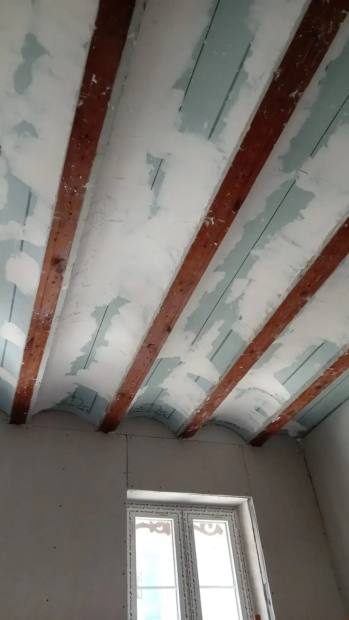 Techo parcialmente terminado con vigas rojas a la vista y aislamiento blanco sobre una ventana.