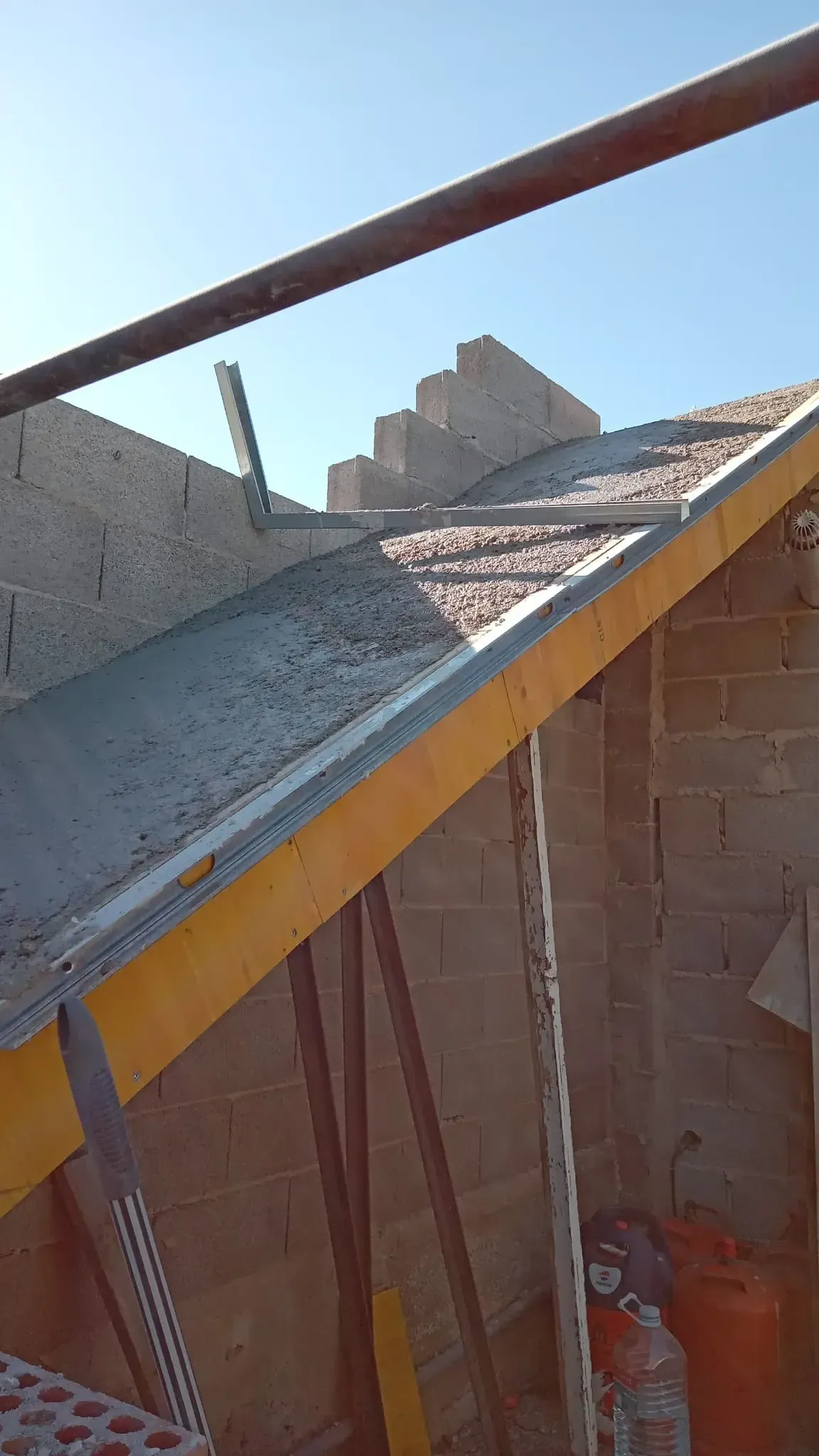 Techo ondulado inclinado en construcción con bloques grises, viga amarilla y barandilla de madera contra el cielo azul.